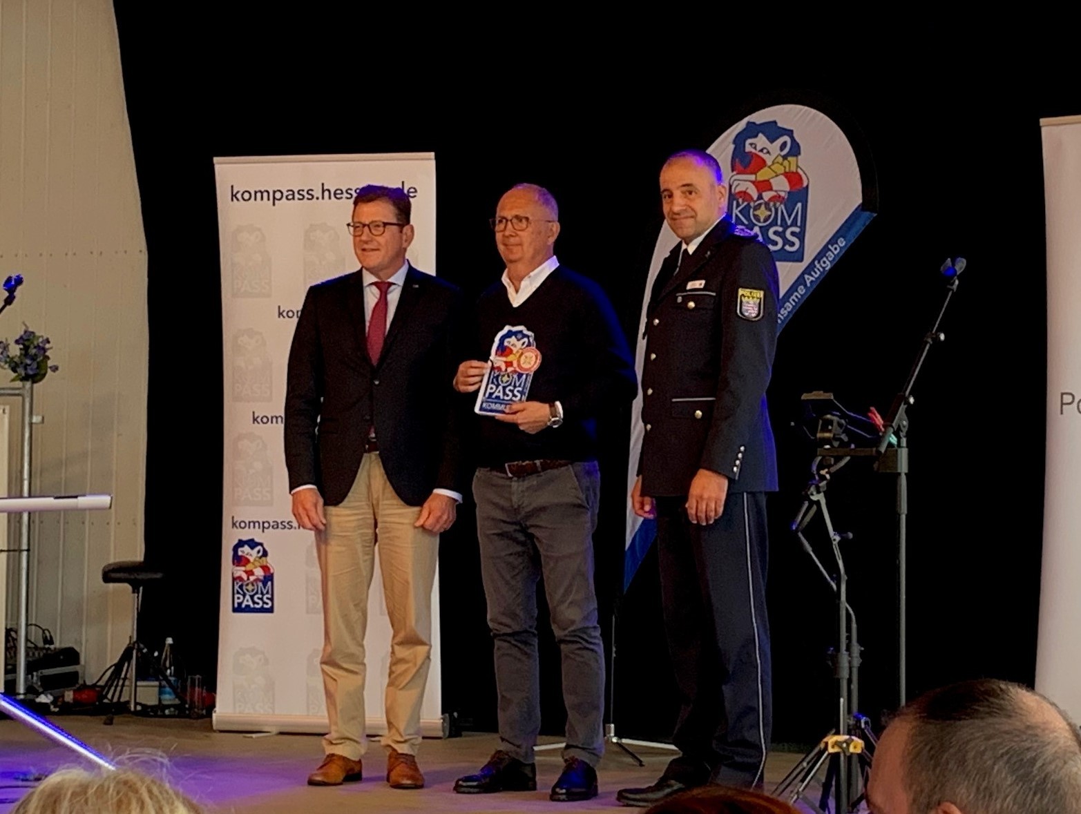Drei Männer auf Bühne, der Mittlere hält Siegel in der Hand