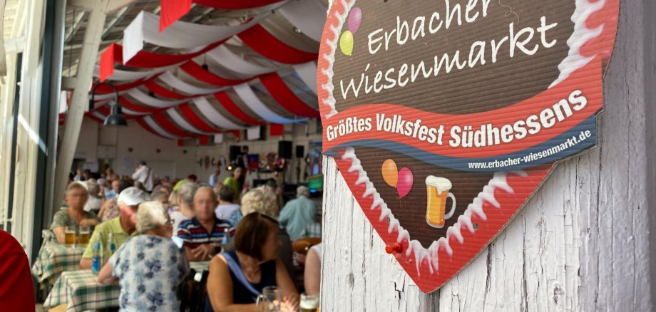 Großes Pappherz mit Aufschrift "Erbacher Wiesenmarkt" im Vordergrund, Menschen an Biertischgarnituren im Hintergrund