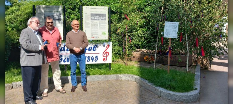 Drei Männer vor Schrifttafel und geschmücktem Brunnen