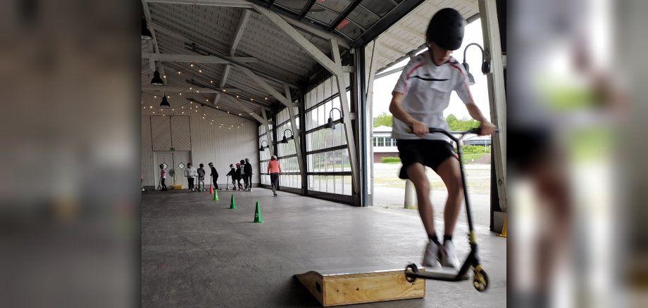 Kind springt mit Scooter über Hindernis. Im Hintergrund sind weitere Kinder beim Scooter-Fahren zu sehen.