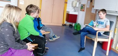 Drei Kita-Kinder hören in Klassenzimmer Schulkind beim Vorlesen zu