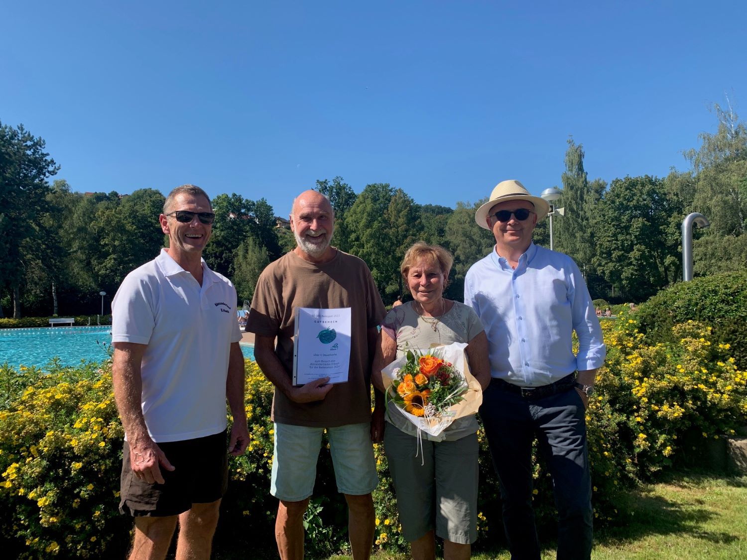 Zwei Männer gratulieren Mann und Frau mit Gutschein und Blumenstrauß im Freibad
