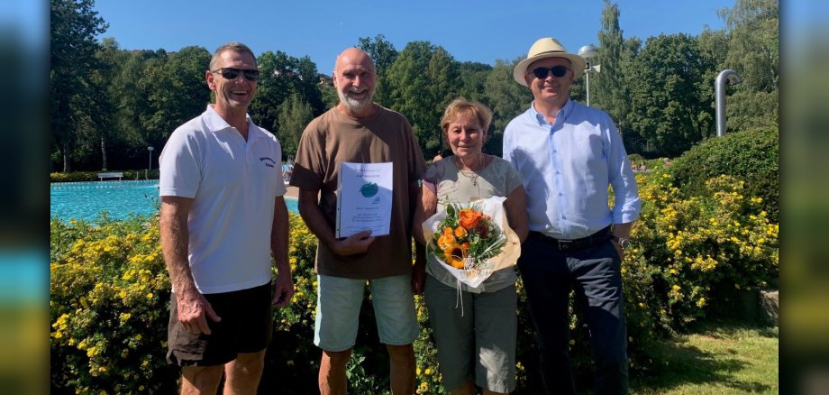 Zwei Männer gratulieren Mann und Frau mit Gutschein und Blumenstrauß im Freibad