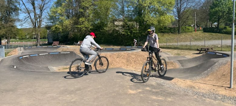 Kinder mit BMX-Bike und Roller warten darauf,  auf einer asphaltierten Pumptrack-Strecke fahren zu können.