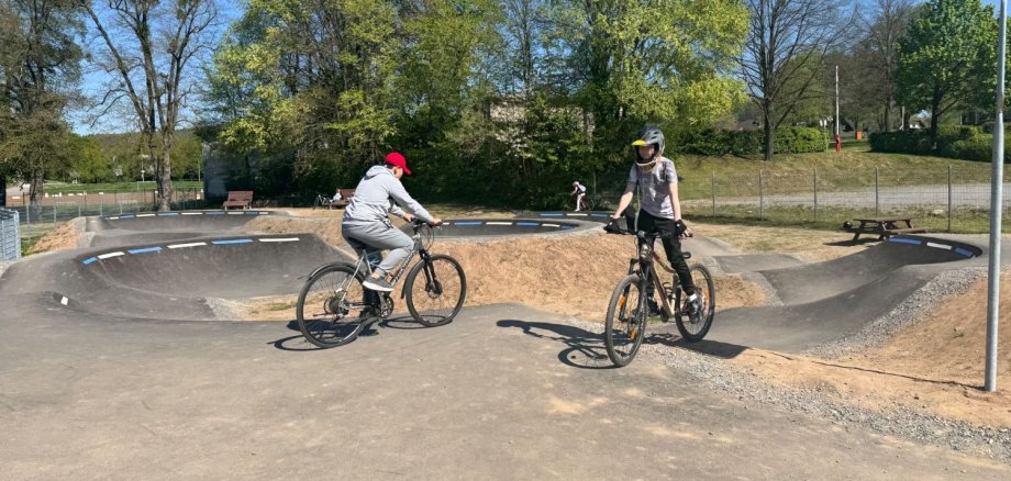 Kinder mit BMX-Bike und Roller warten darauf,  auf einer asphaltierten Pumptrack-Strecke fahren zu können.