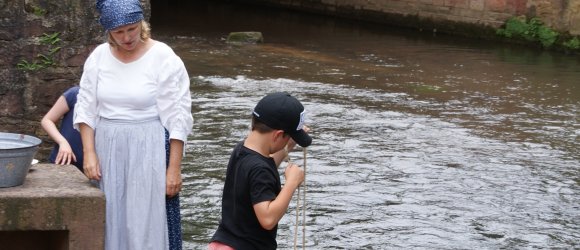 Frau mit historischem Kostüm einer Waschfrau mit Kindern an Flüsschen beim Wäschewaschen