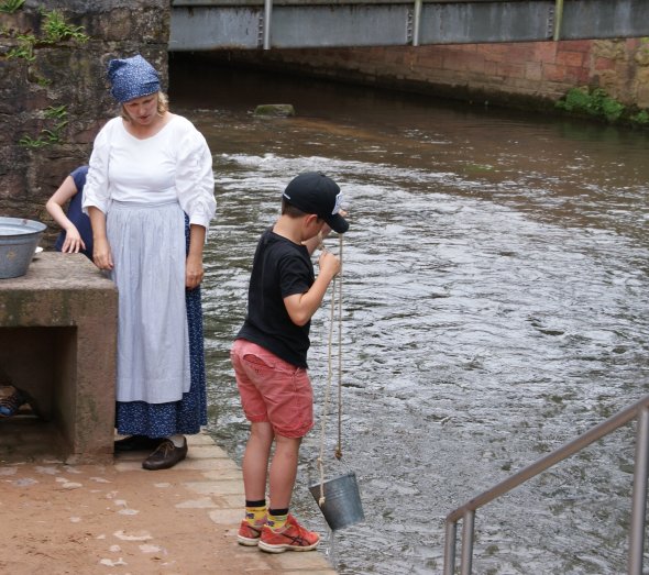 Wäsche waschen mit der Waschfrau Frau mit historischem Kostüm einer Waschfrau mit Kindern an Flüsschen beim Wäschewaschen