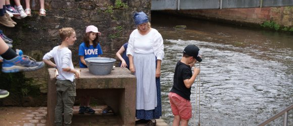 Frau mit historischem Kostüm einer Waschfrau mit Kindern an Flüsschen beim Wäschewaschen